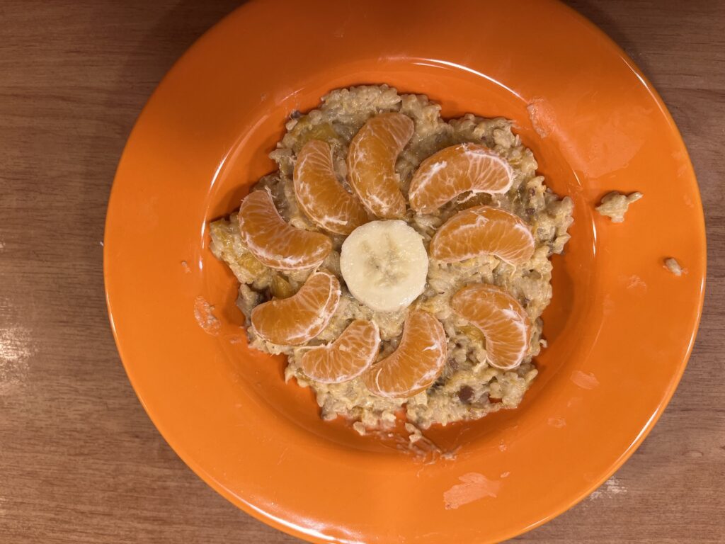 W głębokim, pomarańczowym talerzu, na puddingu ryżowym leżą cząstki banana i mandarynek ułożone w kształt kwiatu.