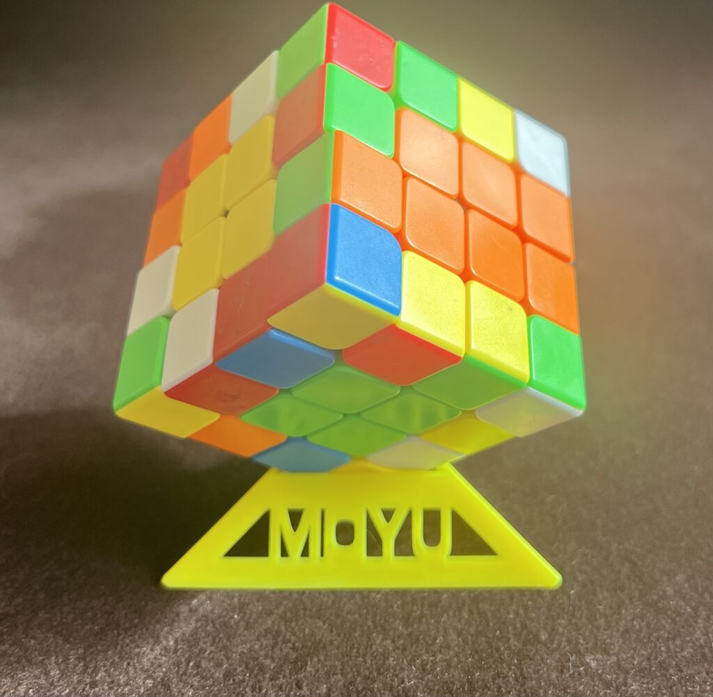 Opis Be My AI: Na zdjęciu widoczna jest kostka Rubika umieszczona na żółtym stojaku. Kostka ma żywe kolory, w tym czerwony, zielony, niebieski, pomarańczowy, biały i żółty. Stojak, na którym stoi kostka, ma kształt trójkąta i jest żółty z napisem "MOYU" w środku. Tło zdjęcia jest w odcieniach szarości.